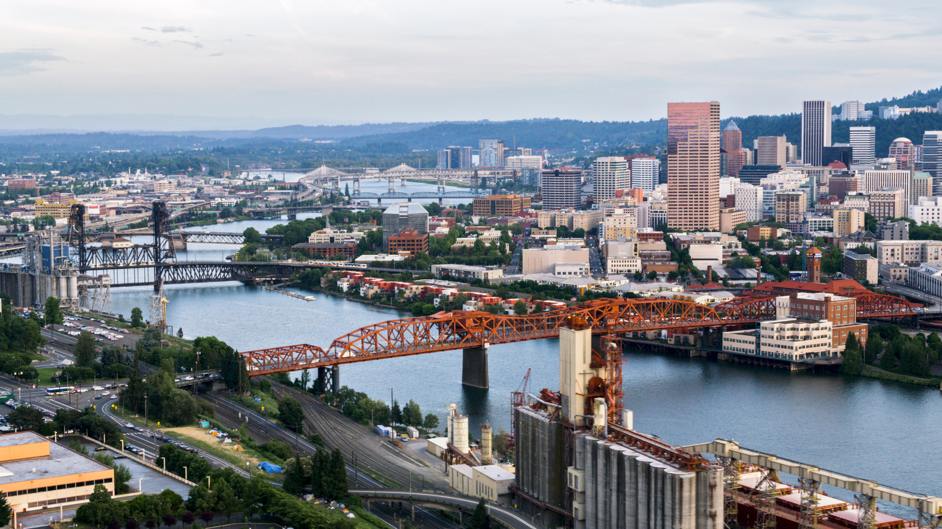 Cityscape of Portland, Oregon, with bridges over the Willamette River, buildings, and a forested hillside in the background.