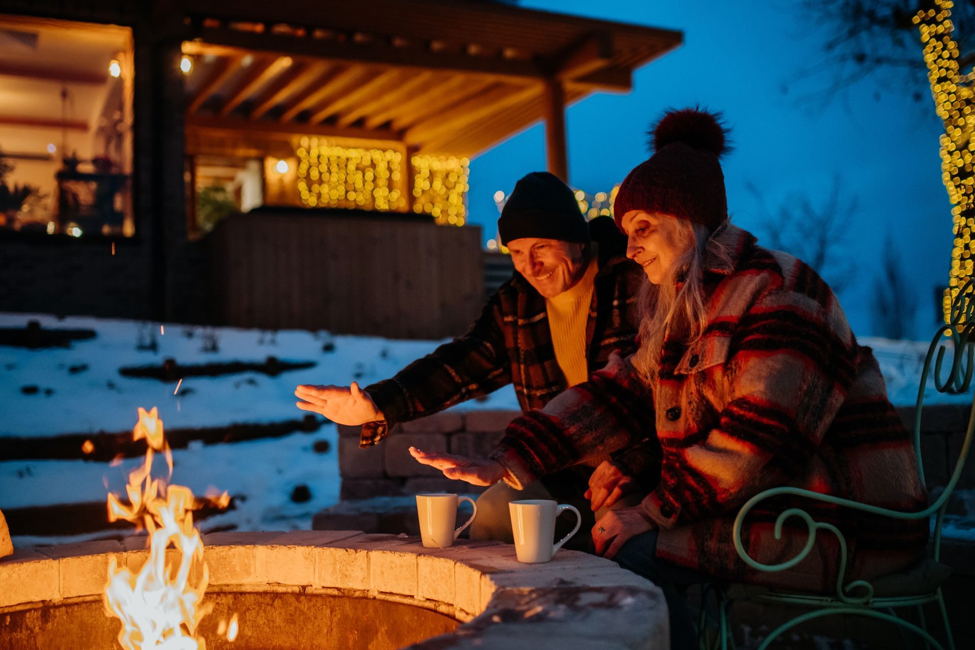 Couple warms hands by a fire pit, mugs nearby, snow-covered landscape at dusk.