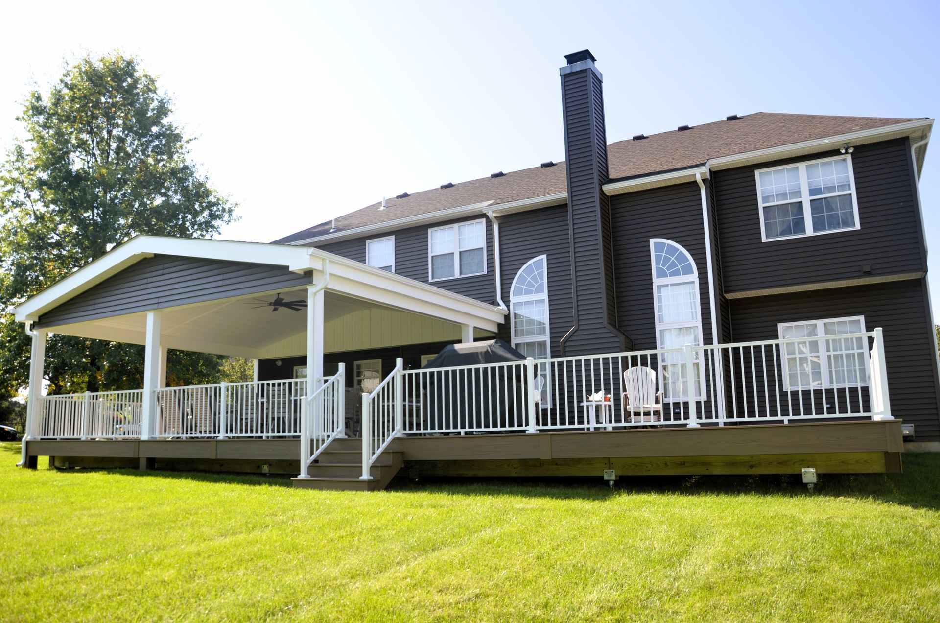 Two-story brown house with white deck and screened porch on a grassy hill