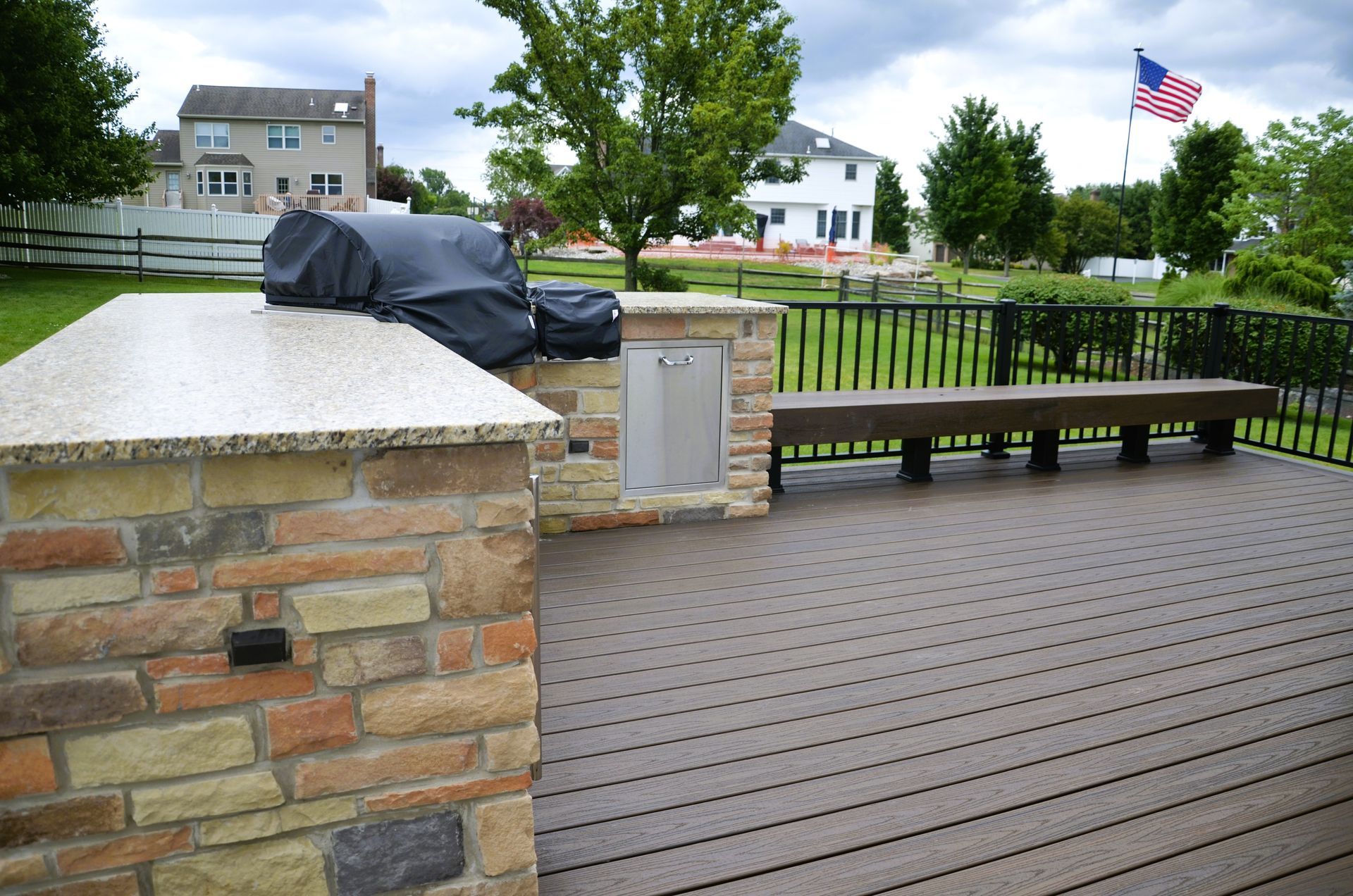 Outdoor kitchen area with grill, trash can, bench, and American flag in background.