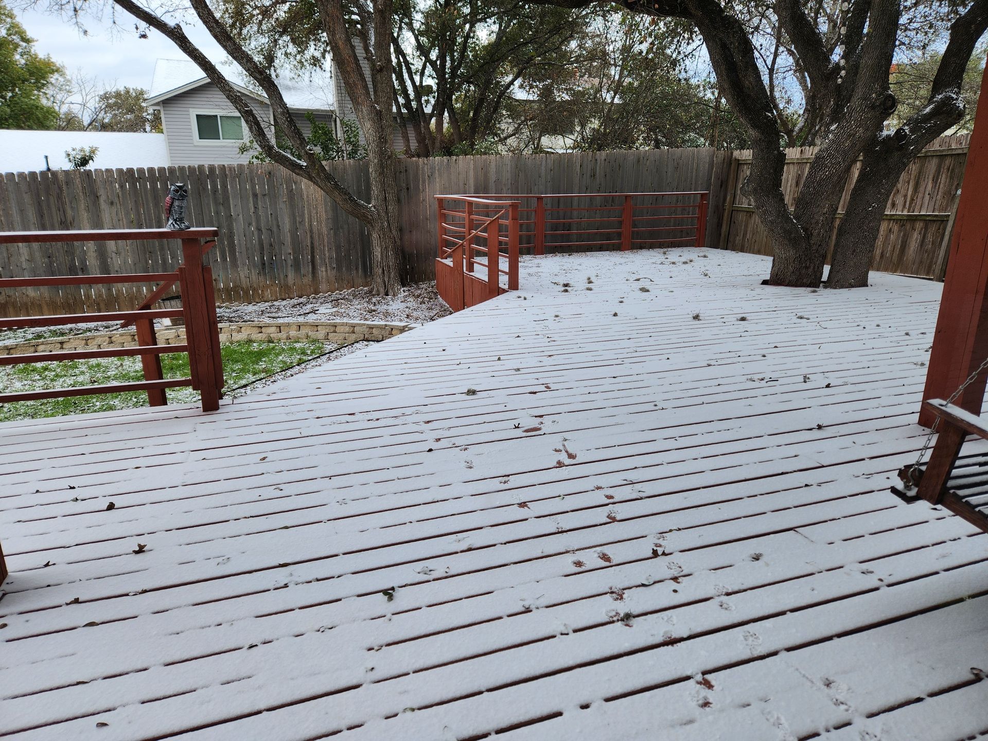 Snow-covered wooden deck in a backyard, surrounded by a fence and trees.