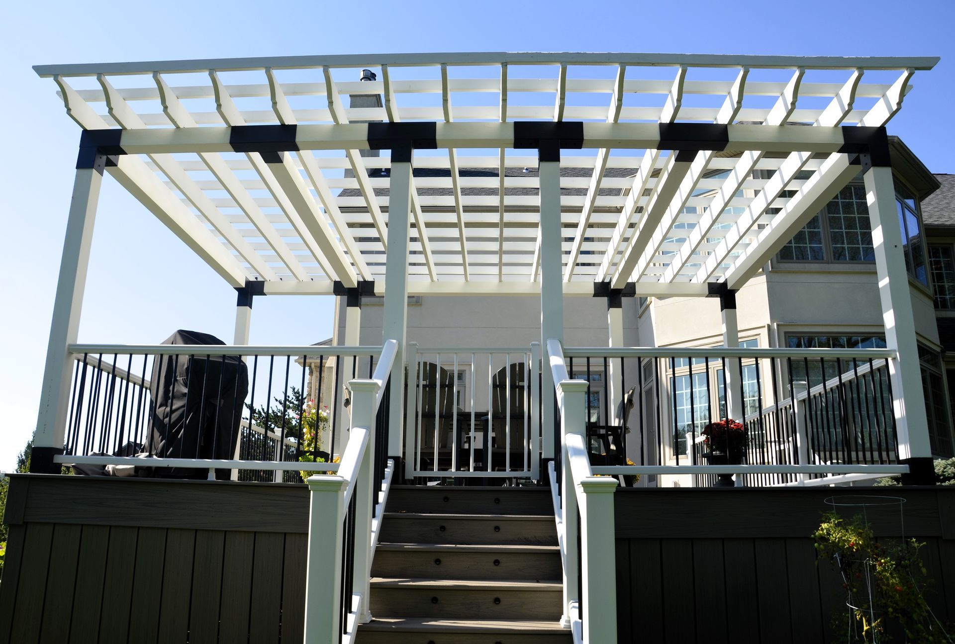 White pergola on a deck with stairs leading up, blue sky in the background.