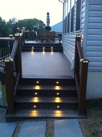 Wooden deck stairs with lit step lights leading to a raised patio beside a house at dusk.