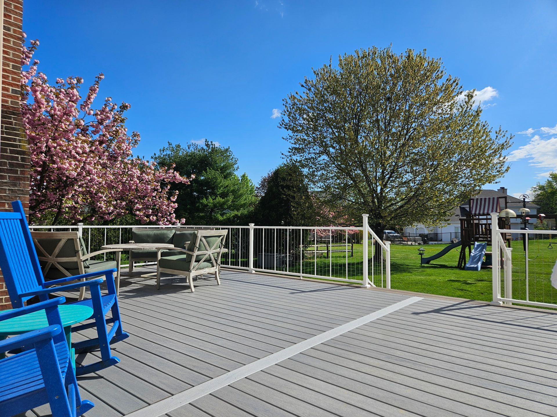 Backyard deck with furniture; lawn, trees, blue sky.