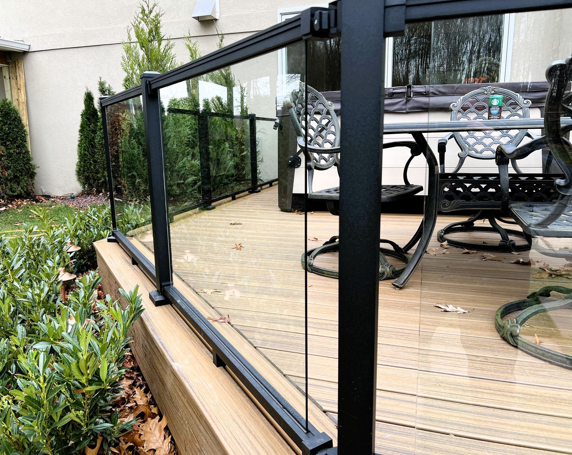 Black metal and glass railing on a wooden deck, with a view of a table and chairs in the background.