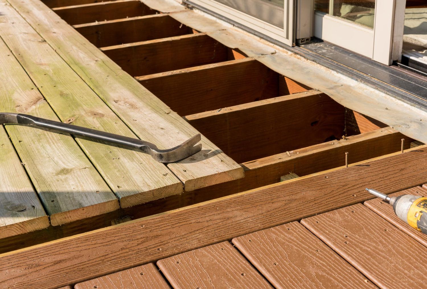 Partially removed deck boards with a pry bar and exposed joists beside a windowed wall