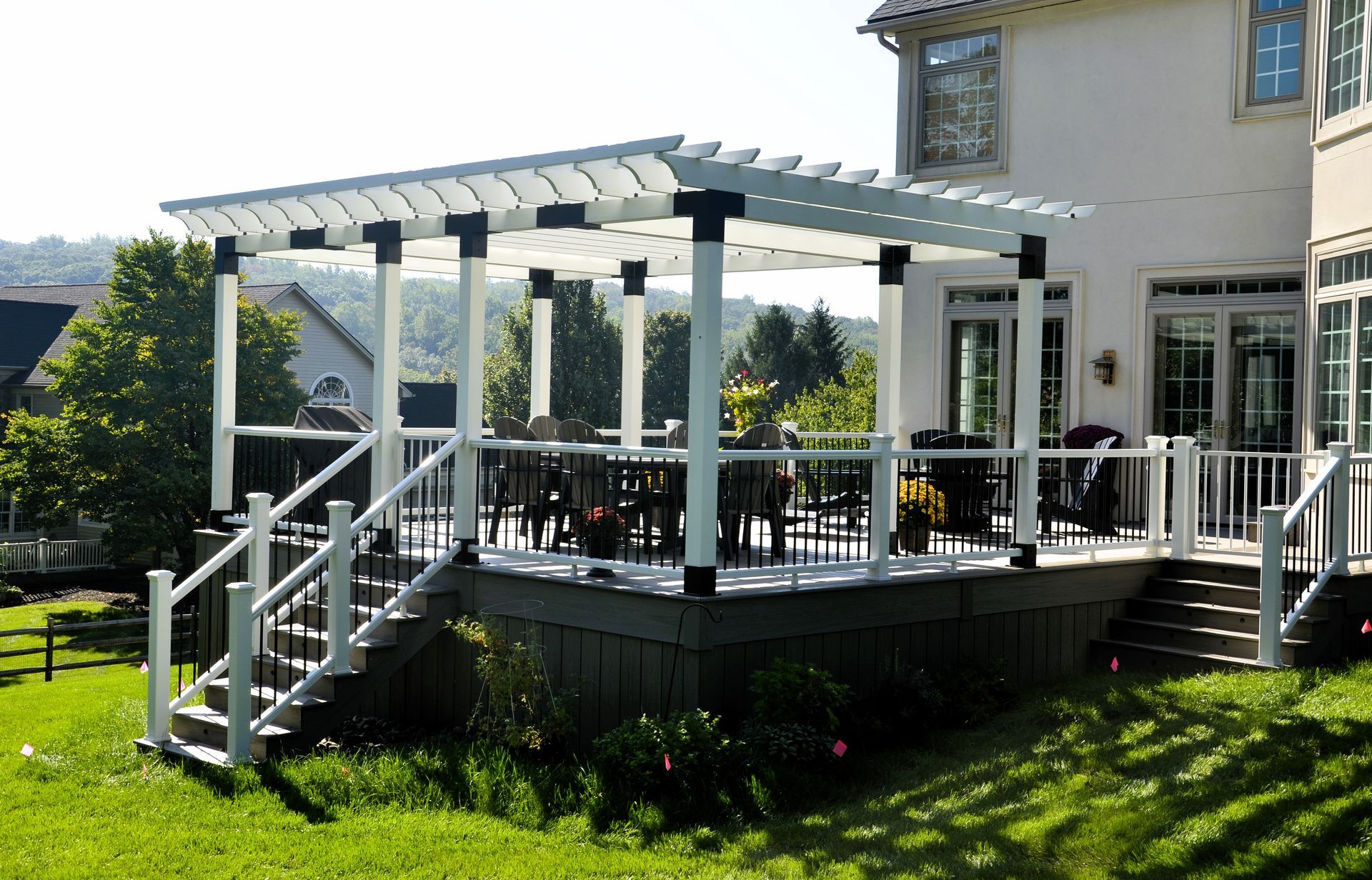 White pergola over deck with dining set, steps down to grassy yard, overlooking trees.