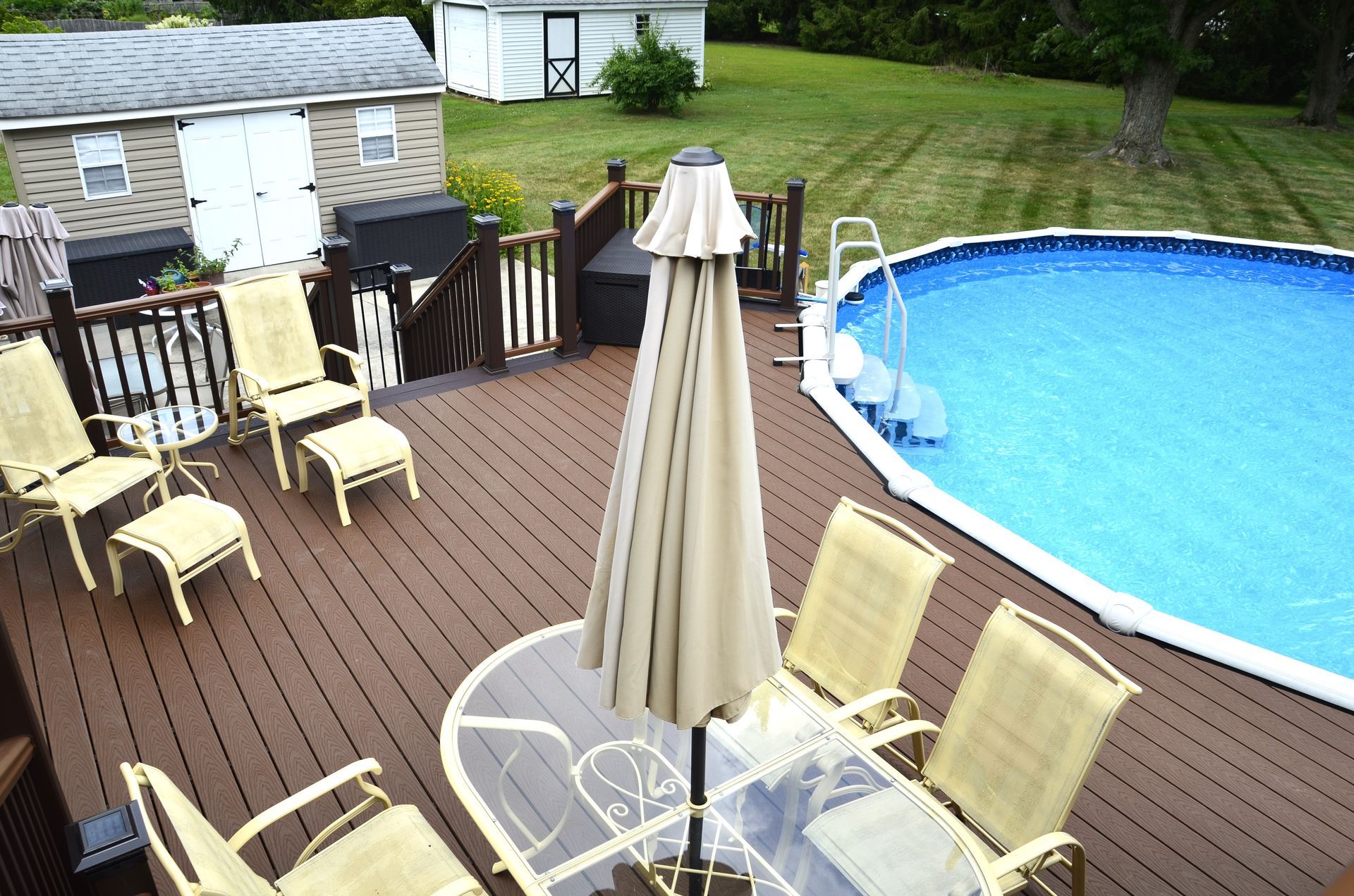 Wooden deck with outdoor furniture next to a pool and lawn with sheds.