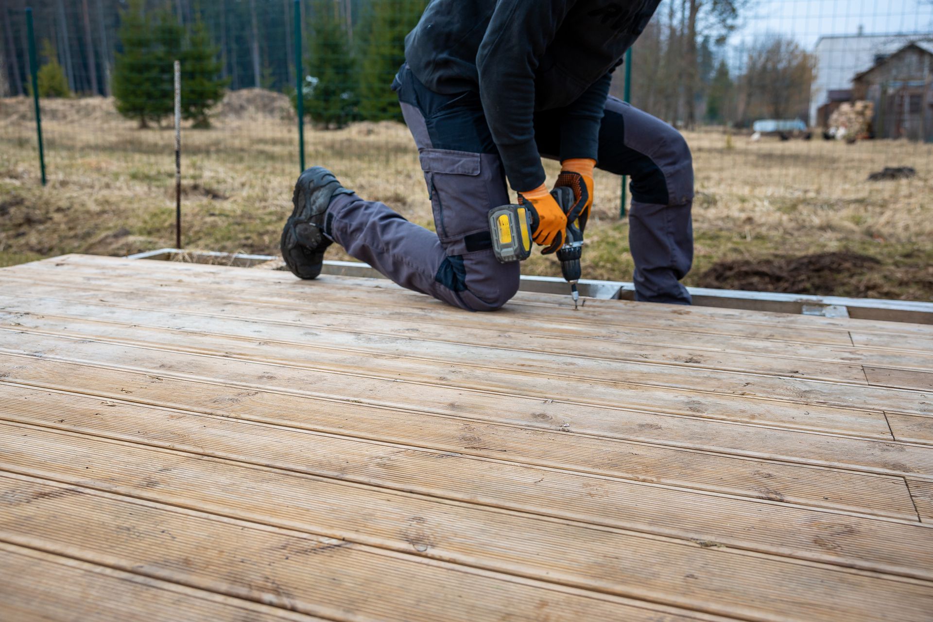 A man is kneeling down while using a drill on a wooden deck.