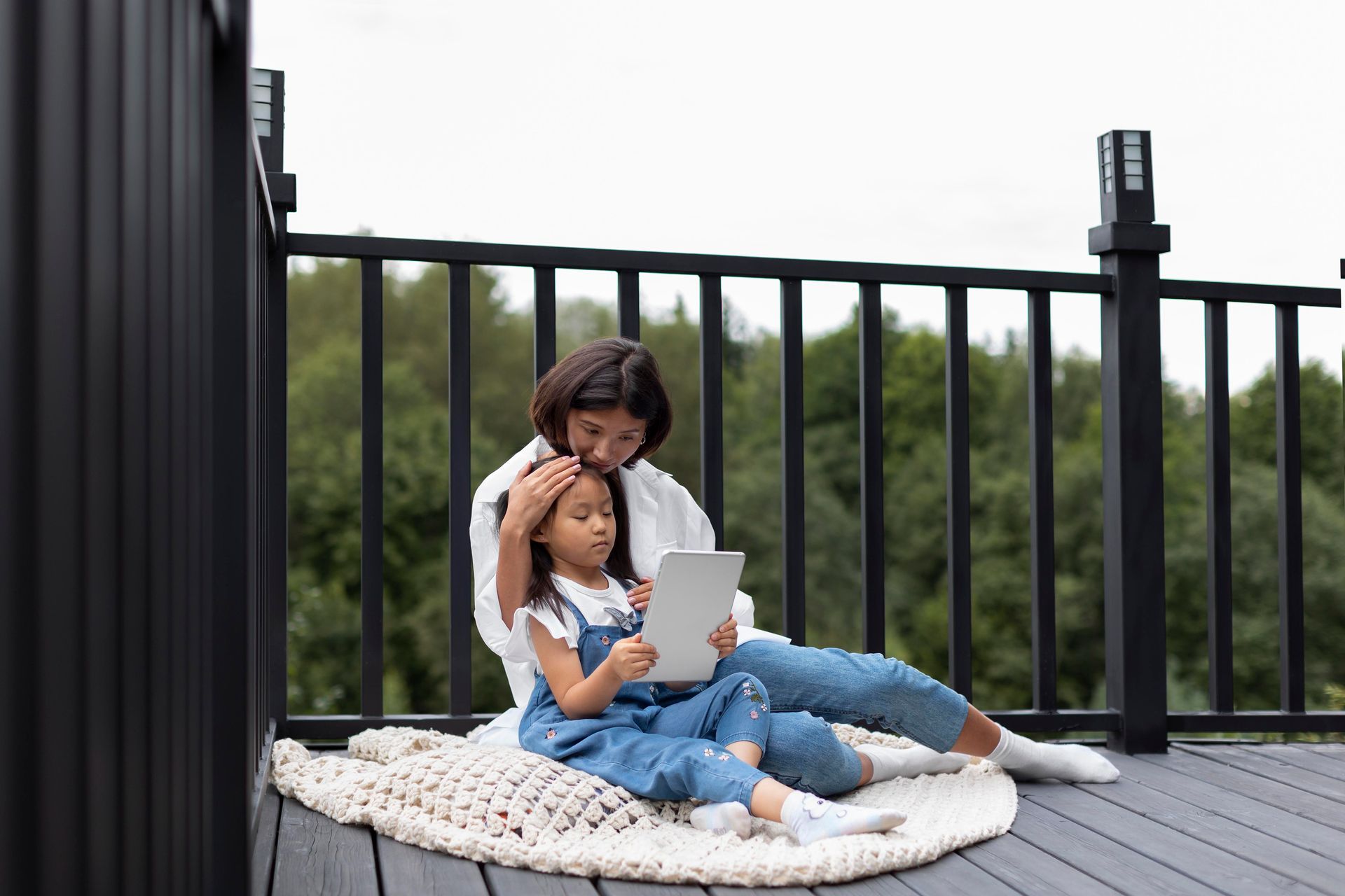 Mother and daughter on deck looking at tablet, wearing casual clothes; greenery background.