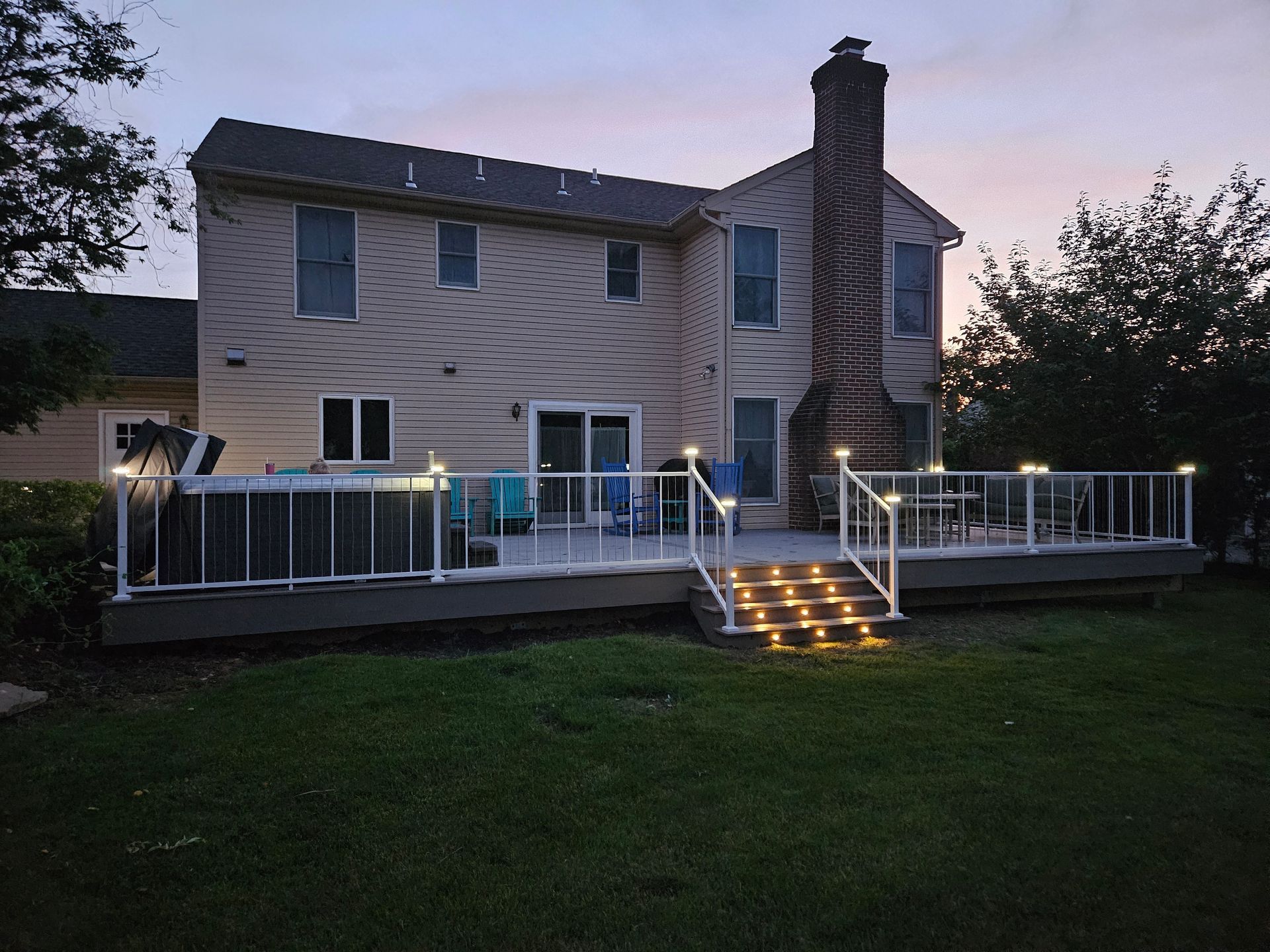 Back view of a two-story beige house with a lit deck and railing. Evening sky.