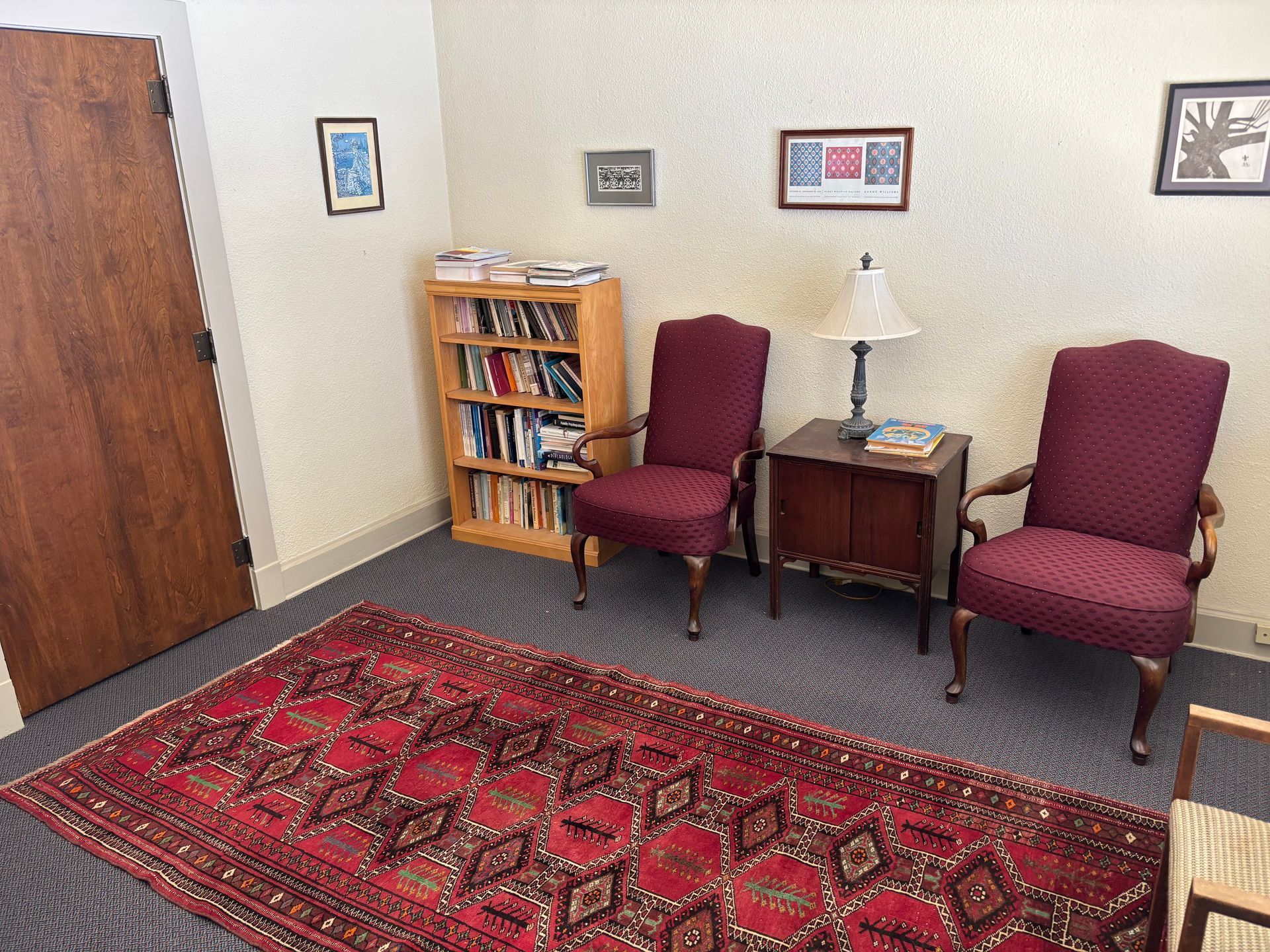 A waiting room with two chairs , a rug , a bookshelf and a lamp.
