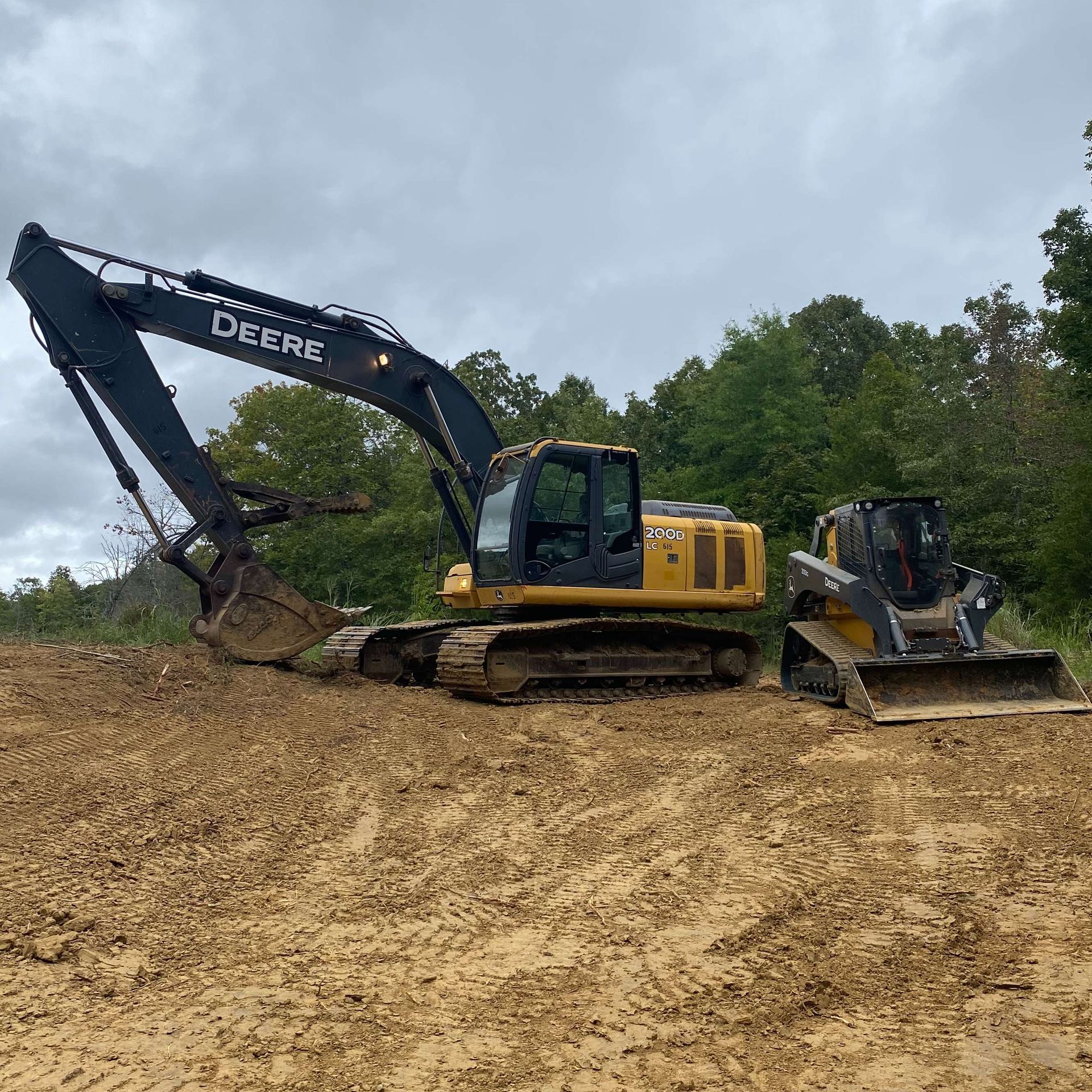 John Deere excavator and skid steer on muddy ground, trees in background under cloudy sky.