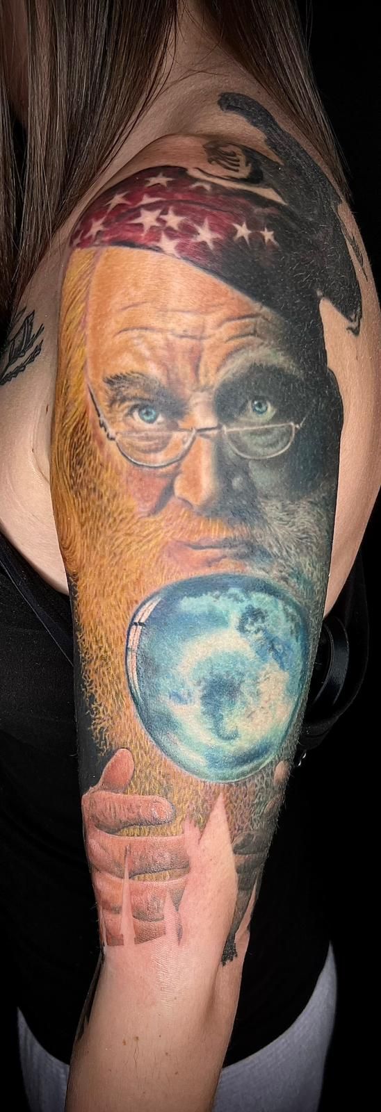 A woman has a tattoo of a man with a beard and glasses on her arm.