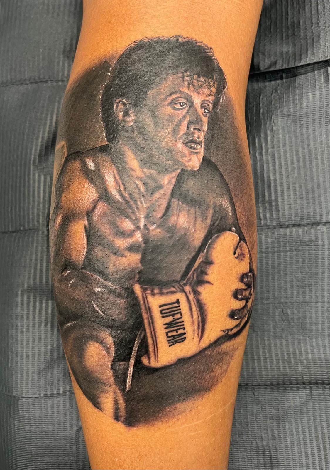 A black and white tattoo of a man wearing boxing gloves on his leg.