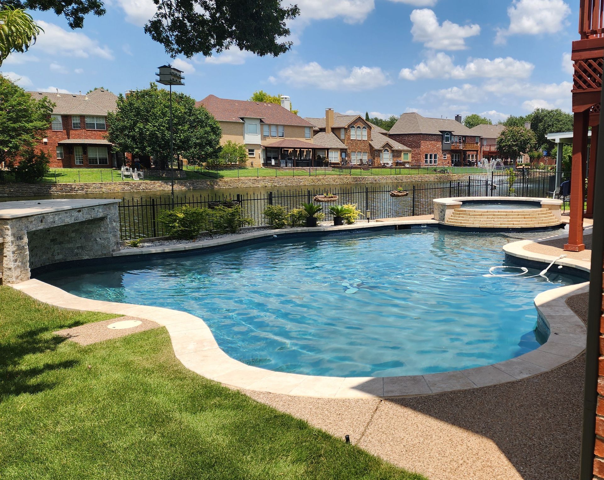 Pool with blue water, surrounded by a patio, lawn, and neighborhood houses under a sunny sky.