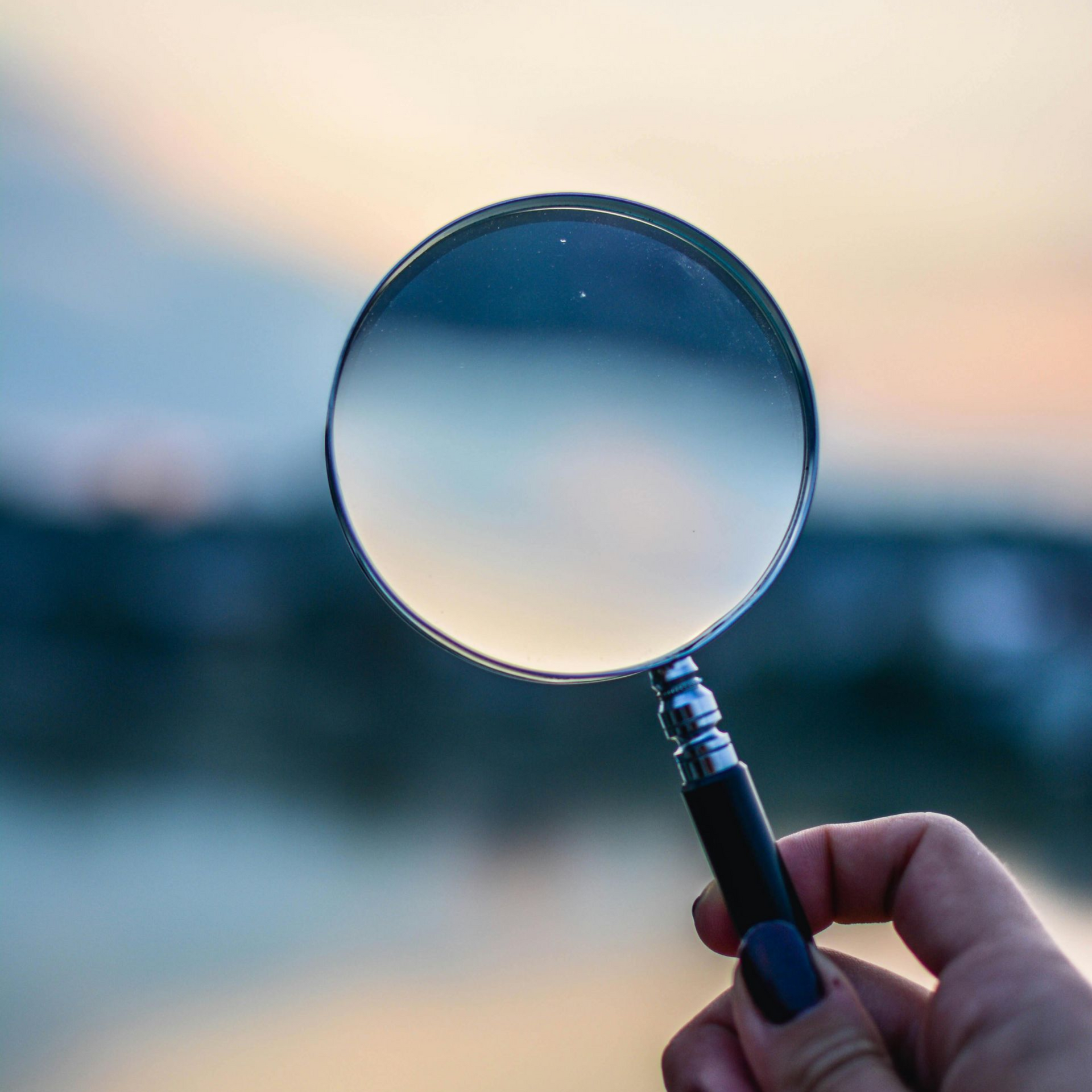 Hand holding a magnifying glass, focusing on a blurry outdoor scene with light blue and orange hues.