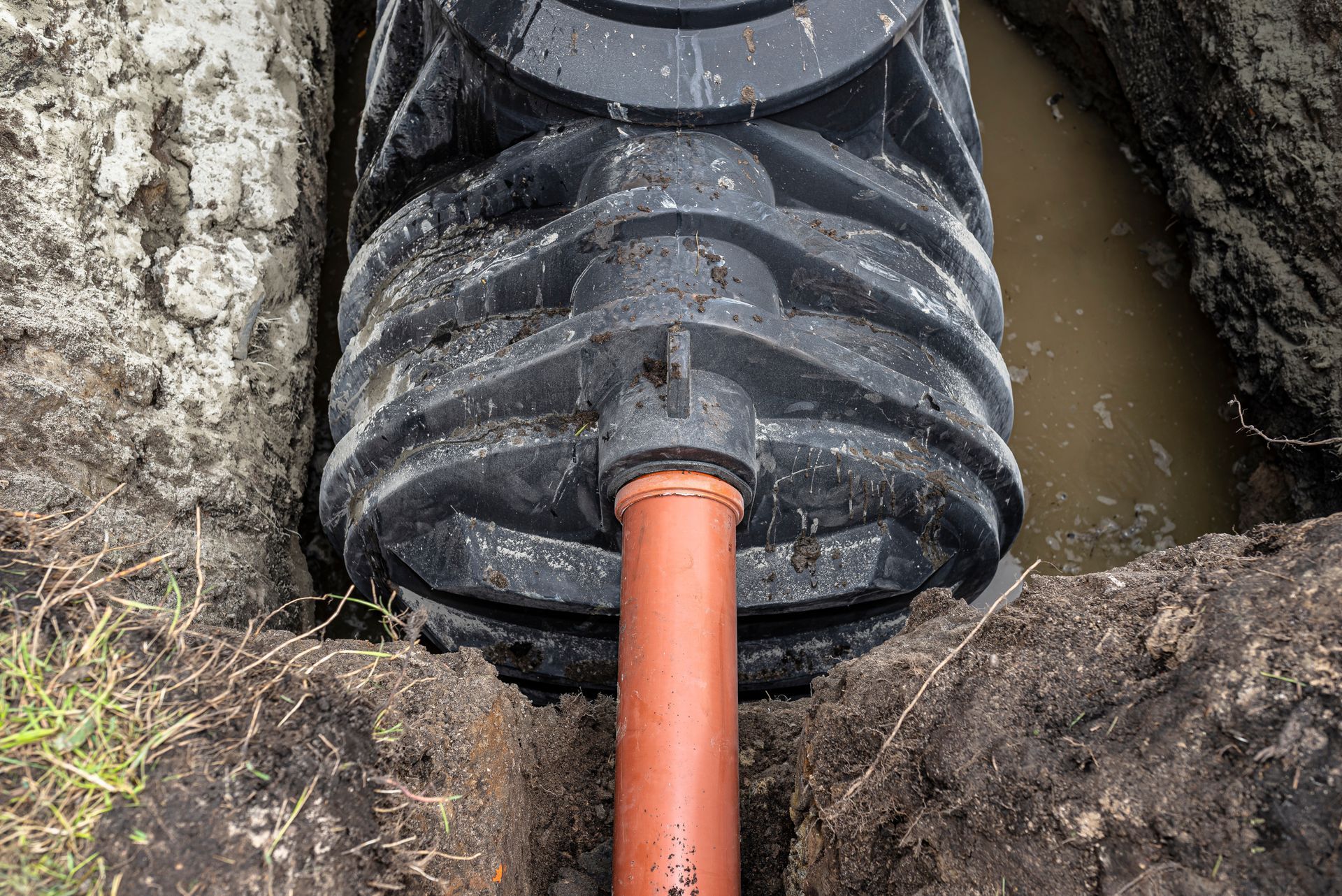 A black plastic drainage tank installed in a muddy trench with an orange PVC pipe connected to its side.