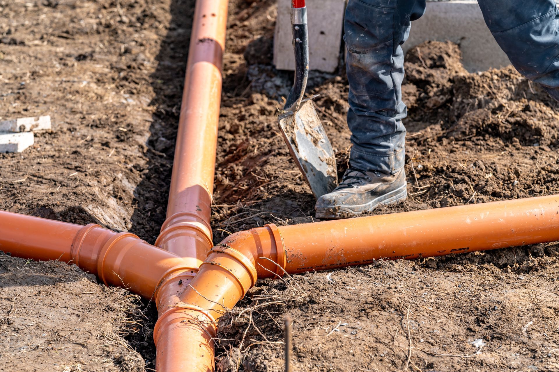 Worker using a shovel to clear dirt around orange PVC drainage pipes laid in a trench.