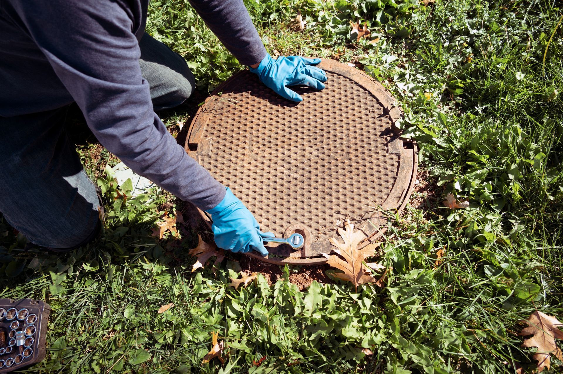 A person wearing blue gloves uses a tool to lift a heavy, circular metal manhole cover set in the grass.