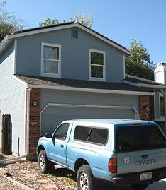 A light blue Toyota pickup truck with a camper shell parked in front of a blue two-story house with a two-car garage.