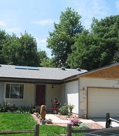A light-colored single-story house with a red door, a front lawn, and a garage under a cloudy blue sky.