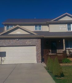 A two-story suburban house with a tan exterior, stone accent on the garage, and a driveway under a clear blue sky.