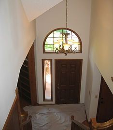 An elevated view of an entryway with a dark wood door, sidelight, arched window, and a chandelier above a drop cloth.