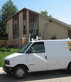 A white service van parked in front of a two-story beige and brown house with multiple ladders leaning against its side.