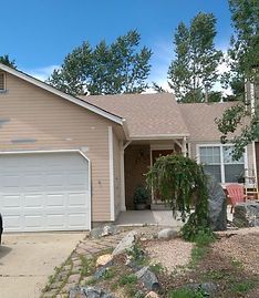 A single-story tan house with a white garage door, surrounded by trees and a stone-bordered front yard.