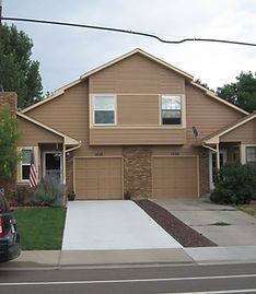A two-story, tan duplex with two attached garages, a concrete driveway, and a small patch of grass.