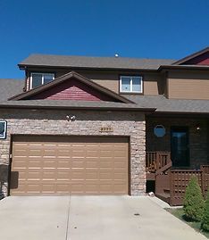 A two-story townhouse with a stone facade, brown garage door, maroon gable, and dark wood stairs under a blue sky.