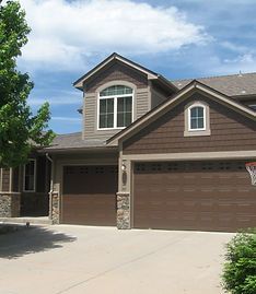 A two-story house with dark brown siding, stone accents, two garage doors, and a concrete driveway under a blue sky.