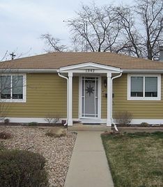 A one-story yellow house with a gabled porch entrance, central front door, and a concrete walkway leading to the house.