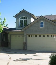 A two-story suburban house with a two-car garage, gray shake siding, and a paved driveway under a clear blue sky.