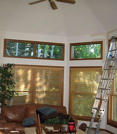 A tall metal ladder leans against the wall of a sunlit living room with large windows, wood blinds, and a leather sofa.