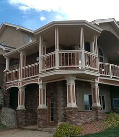 A two-story house exterior featuring a wrap-around balcony with wooden railings and white support columns above stone.