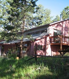 A pink two-story house with a wooden deck nestled in a forest of pine trees under a clear blue sky.
