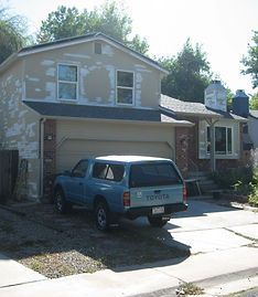 A light brown house with a patchy exterior, a garage, and a light blue Toyota truck parked in the driveway.