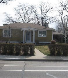 A single-story, tan-sided house with a white front porch entrance, seen from across a street with a sidewalk and hedges.