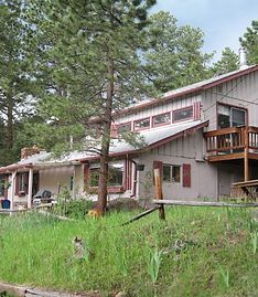A multi-level light-colored house with wooden decks sits on a grassy, pine-covered slope.
