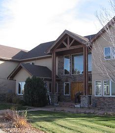 A two-story suburban home with tan stucco walls, stone accents, a dark shingled roof, and a timber-framed entry porch.