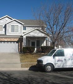 A white work van parked on a suburban street in front of a two-story house with a garage and a tree.