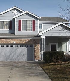A two-story grey suburban house with a two-car garage, brick accents, and a front porch under a clear blue sky.