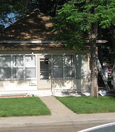 A small, light-colored house with large front windows and a central door sits behind a lawn and a sidewalk.
