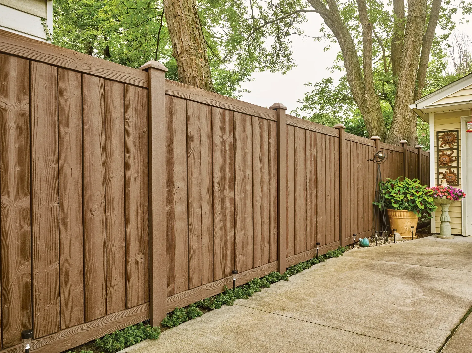 Brown wooden fence bordering a concrete walkway, with a house and greenery in the background.