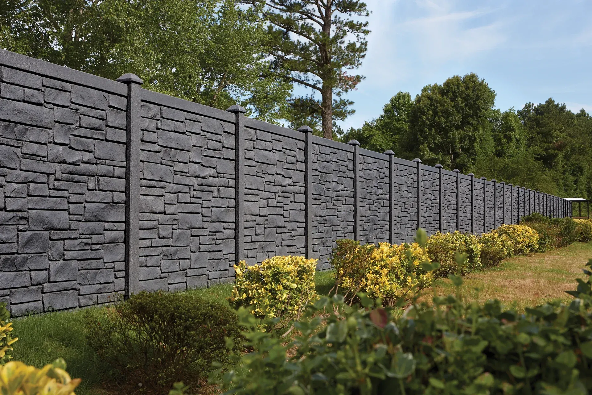 Gray stone-look fence with green bushes in front and trees in the background.