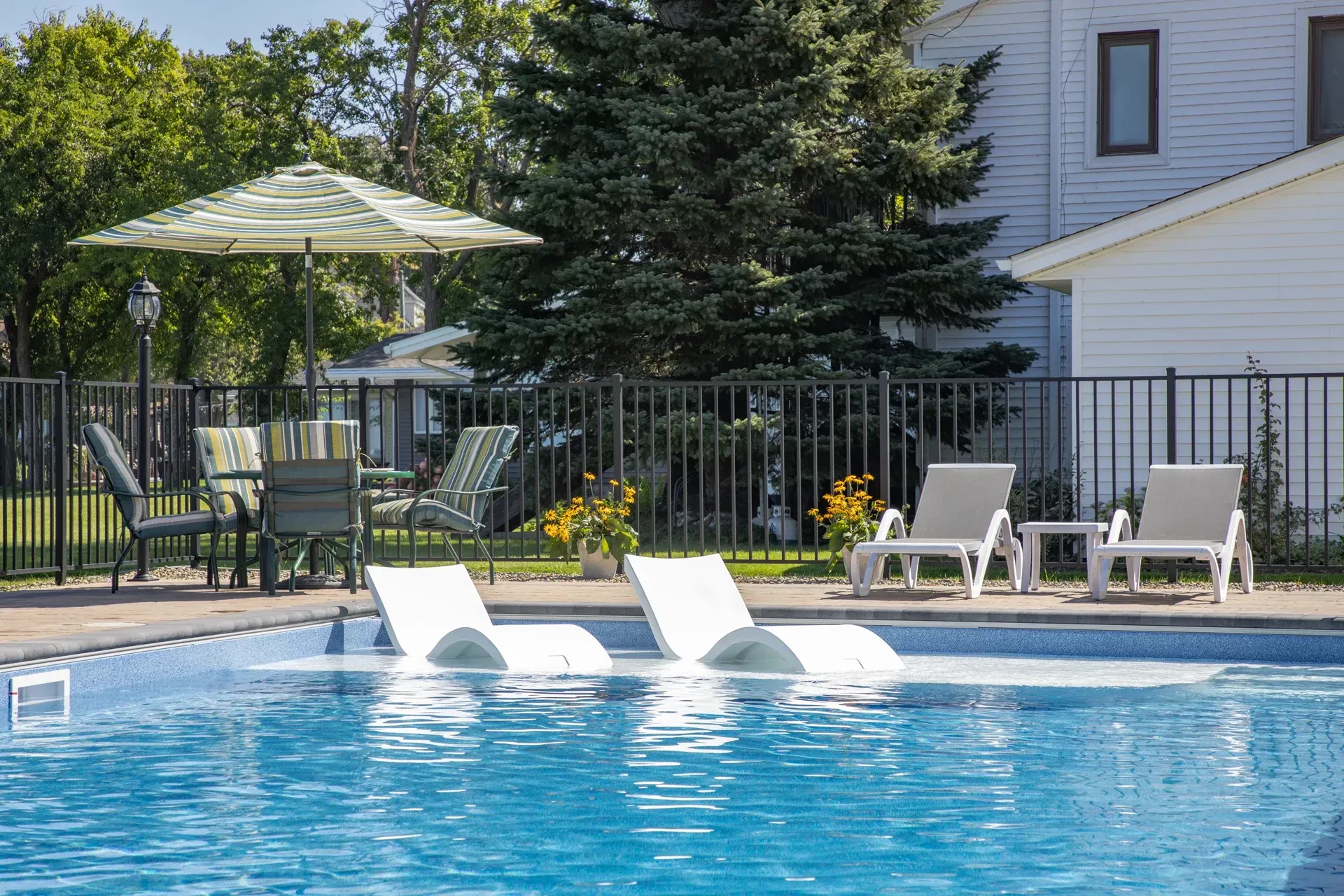 Swimming pool with in-pool loungers, patio furniture, and a black fence, with a building in the background.