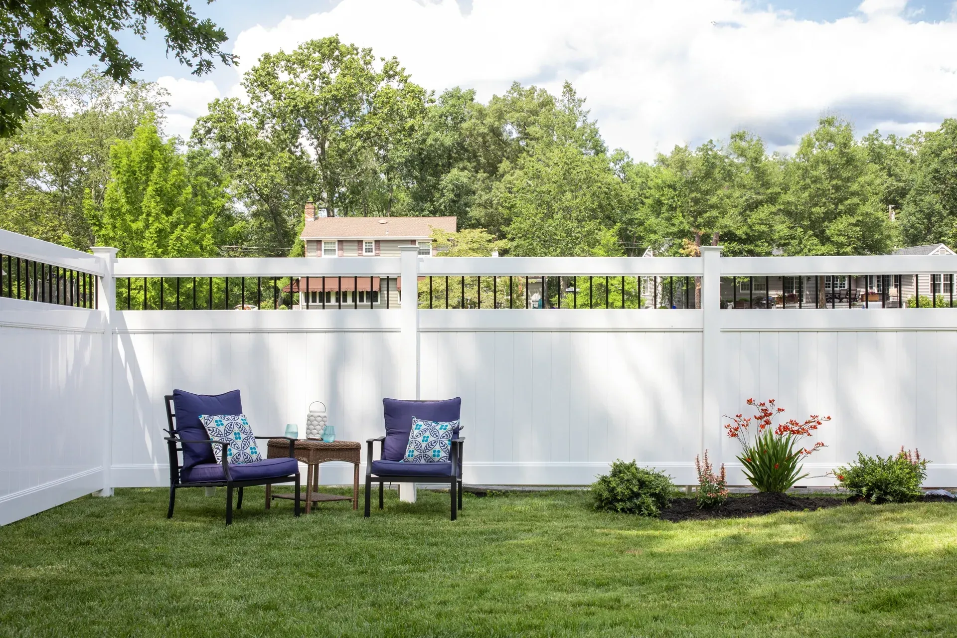 Two blue chairs with pillows sit in a grassy backyard, surrounded by a white fence and greenery.