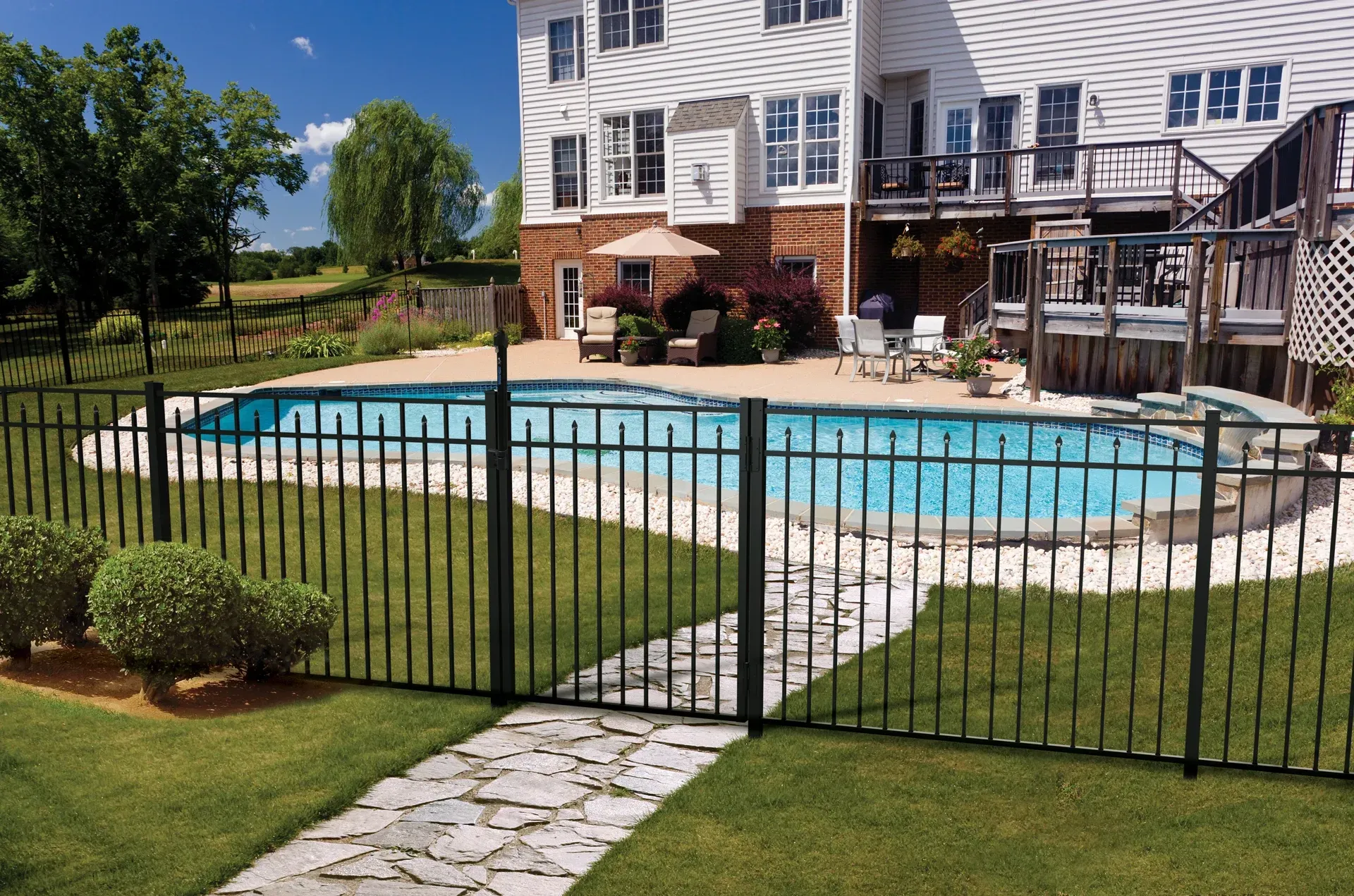 Black fenced swimming pool in a backyard with stone path, patio, and house in the background.
